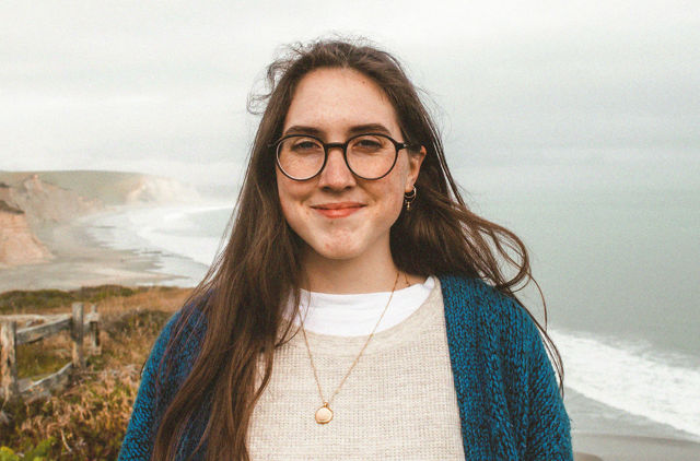 Young female with glasses smiling standing on clifftop with ocean in background
