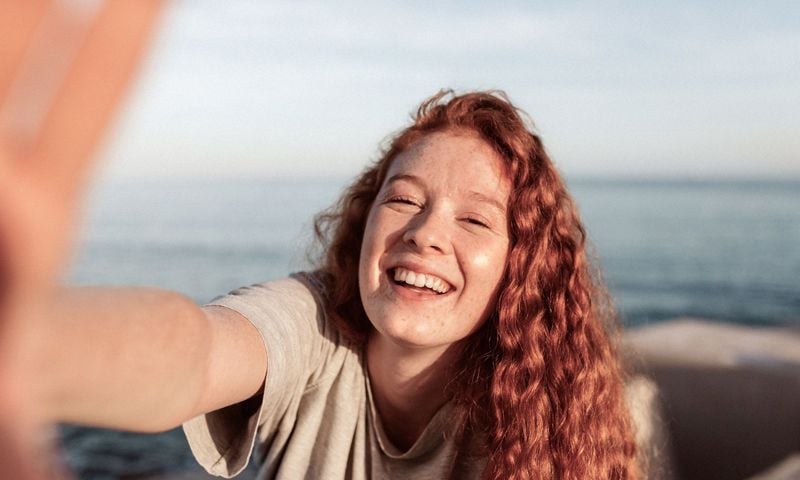 Cheerful young woman with fair skin and curly red hair smiling at the camera while by the sea.