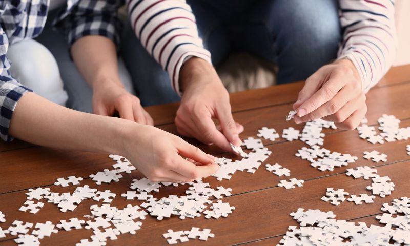 Close up of two people doing a puzzle on a wooden table.