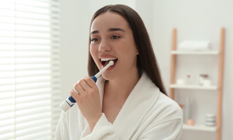 Young woman in a white bathrobe brushing her teeth with an electric toothbrush.